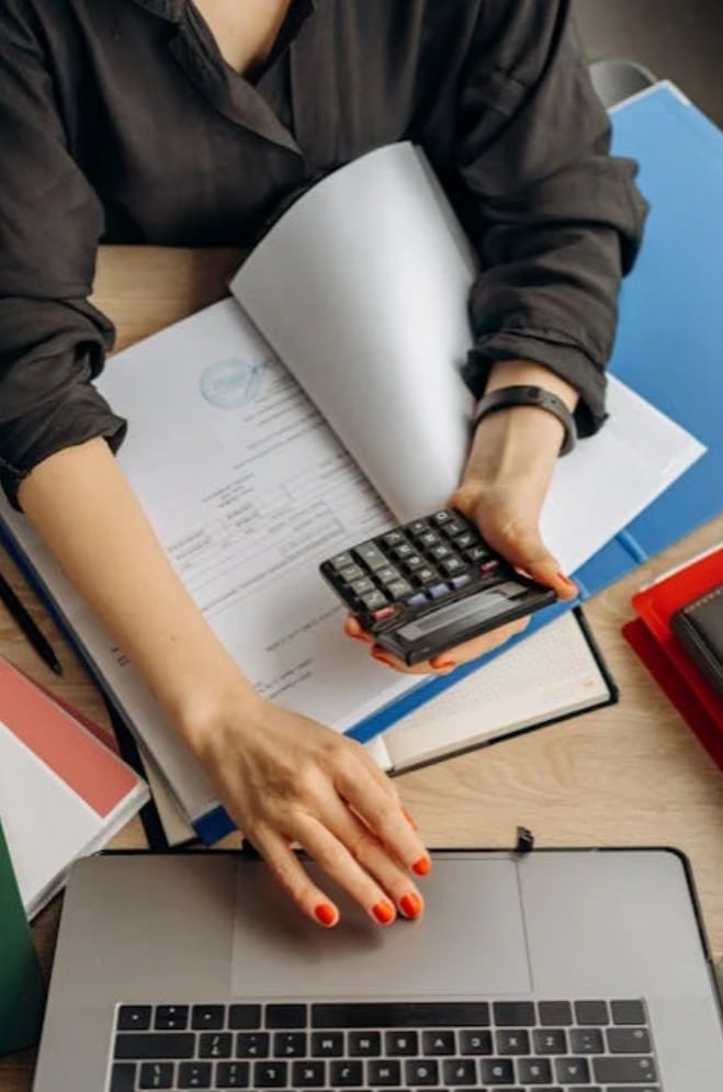 A woman in a black shirt using a calculator and laptop while working with paperwork and folders spread across a desk.